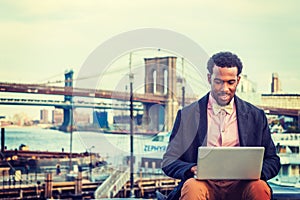 Young black man working on laptop computer ourdoors by river in New York City