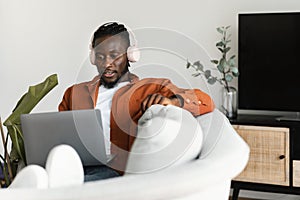 Young black man working on laptop and wearing headphones, sitting on couch at home, free copy space