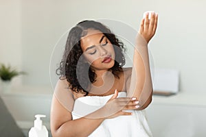 Young black plus size woman in towel applying soothing cream at elbow, moisturizing dry skin at home, free space