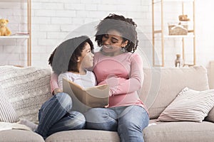 Young black pregnant mother reading book to her daughter at home