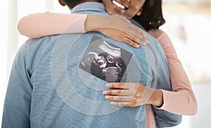 Young black pregnant woman with photo of unborn baby hugging her husband indoors, close up