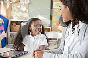 Young black schoolgirl sitting at a table with a tablet computer in an infant school classroom learning one on one with female tea