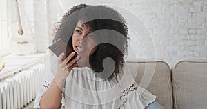 Young black smiling woman recording audio message to friend using smartphone sitting on grey sofa
