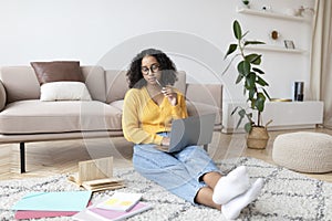 Young black wioman sitting on floor with study materials, having online class with tutor via laptop computer from home