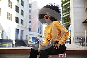 Young African American woman with afro hair sitting on a chair in the street laughing to camera