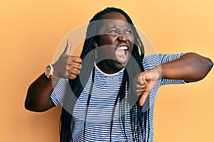 Young black woman with braids doing thumbs down and thumbs up gesture angry and mad screaming frustrated and furious, shouting