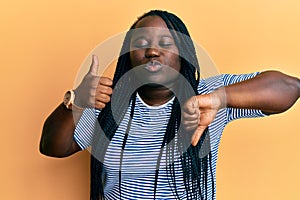 Young black woman with braids doing thumbs down and thumbs up gesture looking at the camera blowing a kiss being lovely and sexy
