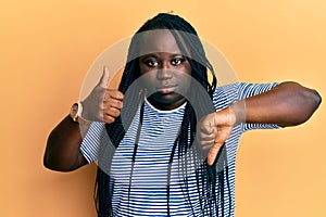 Young black woman with braids doing thumbs down and thumbs up gesture relaxed with serious expression on face