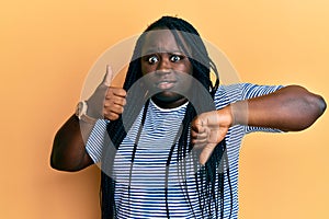 Young black woman with braids doing thumbs down and thumbs up gesture in shock face, looking skeptical and sarcastic, surprised