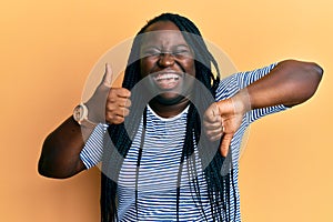 Young black woman with braids doing thumbs down and thumbs up gesture smiling and laughing hard out loud because funny crazy joke