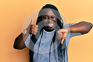 Young black woman with braids doing thumbs down and thumbs up gesture winking looking at the camera with sexy expression, cheerful