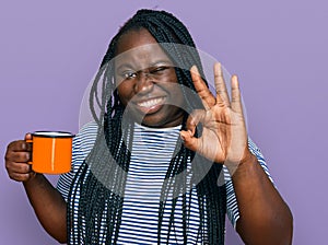 Young black woman with braids drinking a cup coffee doing ok sign with fingers, smiling friendly gesturing excellent symbol