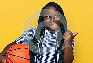 Young black woman with braids holding basketball ball pointing thumb up to the side smiling happy with open mouth