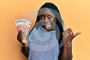 Young black woman with braids holding bunch of 10 euro banknotes pointing thumb up to the side smiling happy with open mouth