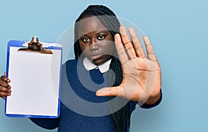 Young black woman with braids holding clipboard with blank space with open hand doing stop sign with serious and confident
