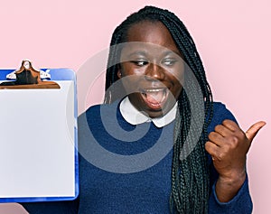 Young black woman with braids holding clipboard with blank space pointing thumb up to the side smiling happy with open mouth