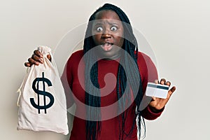 Young black woman with braids holding dollars bag and credit card afraid and shocked with surprise and amazed expression, fear and