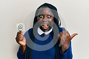Young black woman with braids holding email symbol on paper pointing thumb up to the side smiling happy with open mouth
