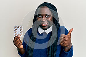 Young black woman with braids holding pills smiling happy and positive, thumb up doing excellent and approval sign