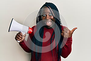 Young black woman with braids shouting through megaphone pointing thumb up to the side smiling happy with open mouth