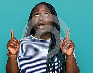 Young black woman with braids wearing casual clothes amazed and surprised looking up and pointing with fingers and raised arms