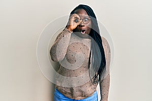 Young black woman with braids wearing casual clothes and glasses doing ok gesture shocked with surprised face, eye looking through