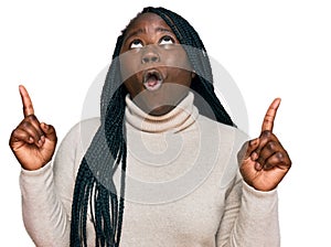 Young black woman with braids wearing casual winter sweater amazed and surprised looking up and pointing with fingers and raised