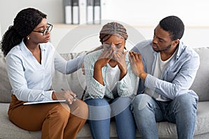 Young black woman crying at therapist& x27;s office, affectionate husband supporting her with professional psychologist