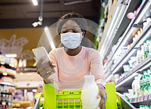 Young black woman in face mask looking into her grocery smartphone app while shopping at dairy section of supermarket