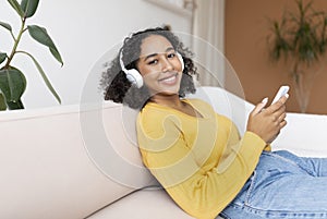 Young black woman in headphones relaxing on couch, listening to music or watching movie on cellphone at home