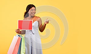 Young black woman holding shopping bags and empty sign with mockup, pointing at blank space over orange background