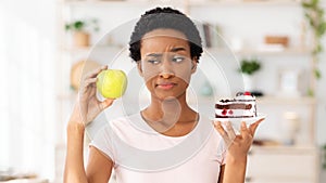 Young black woman looking at apple with suspicion, not wanting to eat fruit instead of cake at home