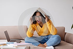 Young black woman shouting with book on her head near laptop computer at home, copy space