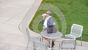 A young black woman is sitting on a chair by a green lawn, drinking coffee, taking a break
