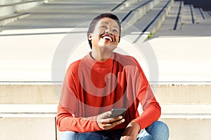 Young black woman sitting outside with cellphone and laughing while looking up