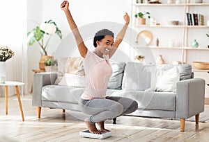 Young black woman sitting on scales, raising hands up in excitement, happy with result of her weight loss diet at home