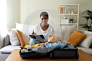 Young Black woman sitting on sofa checking packing list before summer holidays. Preparing suitcase for vacations.