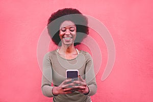 Young black woman standing over pink background using smart mobile phone - African girl laughing and smiling using web app on
