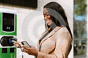 Young black woman using mobile phone while charging her electric car