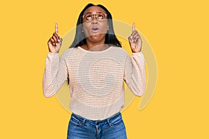 Young black woman wearing casual clothes and glasses amazed and surprised looking up and pointing with fingers and raised arms