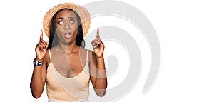 Young black woman wearing summer hat amazed and surprised looking up and pointing with fingers and raised arms