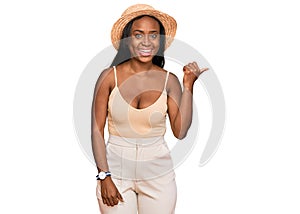 Young black woman wearing summer hat smiling with happy face looking and pointing to the side with thumb up