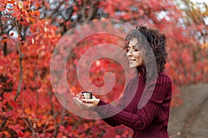 Young blackhaired woman in red sweather with cup of hot drink in red autumnn bushes