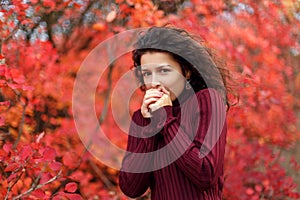 Young blackhaired woman in red sweather heating her hands by blowing stay in red autumnn bushes
