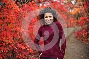 Young blackhaired woman in red sweather posing on camera in red autumnn bushes