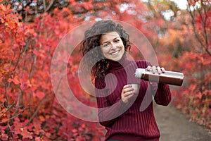 Young blackhaired woman in red sweather with thermos in red autumnn bushes
