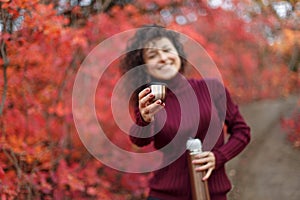 Young blackhaired woman in red sweather with thermos in red autumnn bushes offers to drink a cup of hot drink