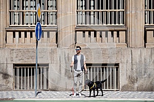 Young blind man with white cane and guide dog standing on pavement in city.