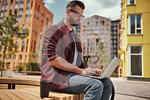 Young blogger. Handsome man with stubble in casual clothes and eyeglasses working on laptop while sitting on the bench
