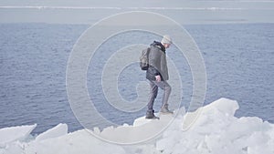 Young blond bearded handsome man wearing warm jacket, hat and boots climbing the glacier, walking on the edge. Amazing